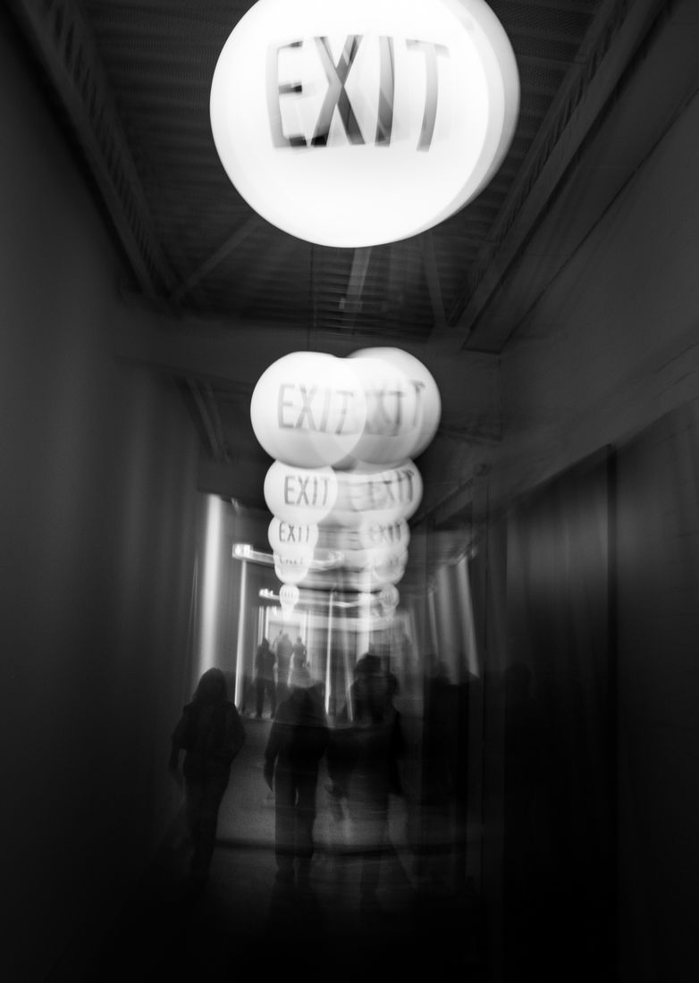 A row of glowing EXIT signs repeated in motion blur above people walking down a corridor