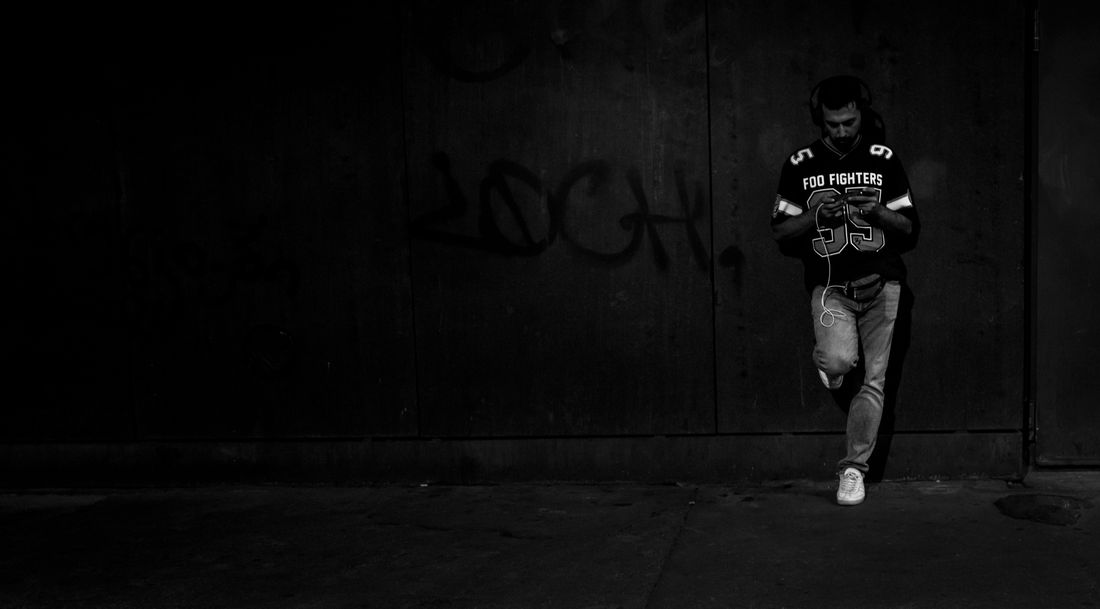 A man in headphones leans against a dark wall, looking at his phone